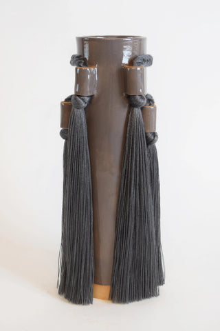 A tall, cylindrical ceramic vase in a dark brown glossy glaze, decorated with two dark gray tassels hanging from wrapped bands near the top and a small light wood base, photographed against a plain white background.