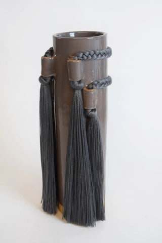 A dark brown ceramic cylindrical vase with a glossy finish, decorated with dark gray braided cords and matching tassels, set against a plain off-white background.