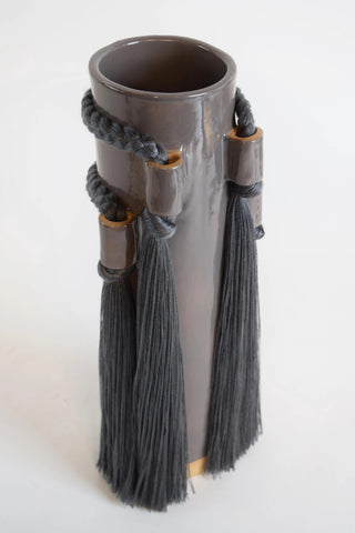 A dark brown ceramic cylindrical vase with a glossy finish, decorated with dark gray braided cords and matching tassels, set against a plain off-white background.
