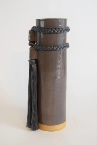 A dark brown cylindrical ceramic vase with a light tan base, wrapped with a dark gray braided leather strap that features a tassel, photographed against a plain off-white background.