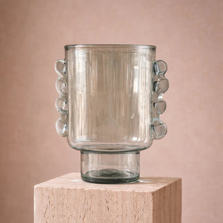 Clear glass vase with bubble-like handles on a wooden block against a pink background