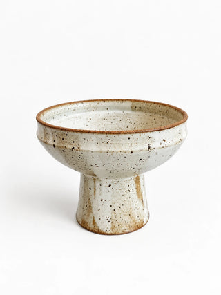 A handmade ceramic pedestal bowl with a speckled off-white glaze and a rustic brown rim, photographed on a clean white background.