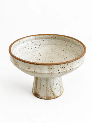 A handmade ceramic pedestal bowl with a speckled cream glaze and a rustic brown rim, set against a plain white background.