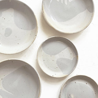 Five round ceramic plates with a speckled light gray and off-white glaze, arranged on a white textured background.