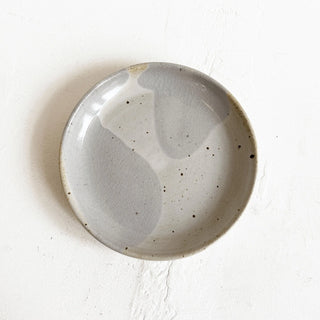 A round handmade ceramic plate with a matte, speckled finish in light gray and off-white tones