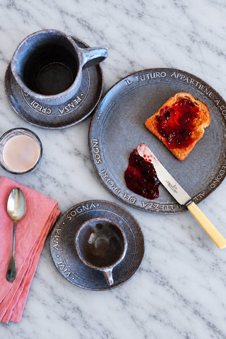 A styled flat-lay of handmade dark gray ceramic tableware on a white marble surface, featuring a plate with toast and jam, cups, a pink napkin, and a spoon.