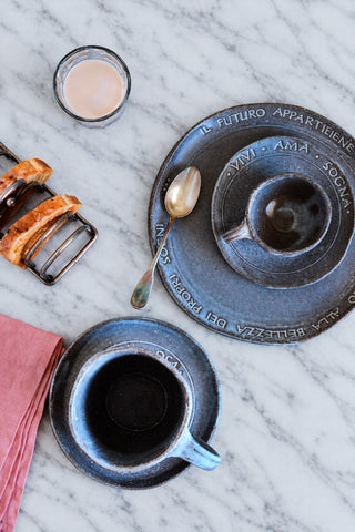 Two dark blue-gray ceramic espresso cups with matching saucers are arranged on a white marble surface, alongside a spoon, a small glass of pale pink beverage, and a pink napkin. The cups have embossed Italian text around the rim.