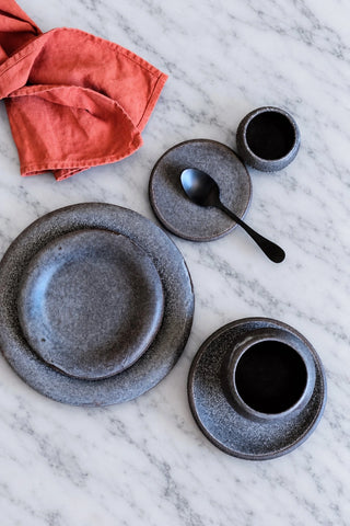 A flat-lay of a dark gray speckled ceramic tableware set on a white marble surface, including plates, a cup, a spoon, and a folded coral-orange cloth napkin.