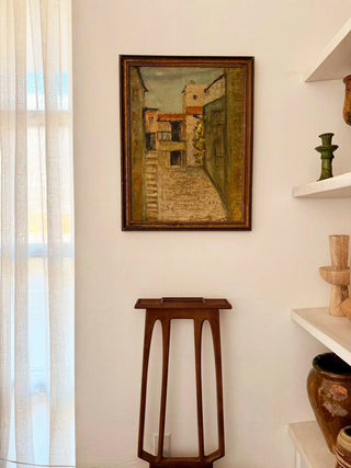 Framed artwork on a wall next to a wooden stool and shelves with decorative items.
