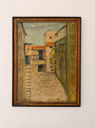 Framed painting of a street scene with buildings and a tree on a white wall.
