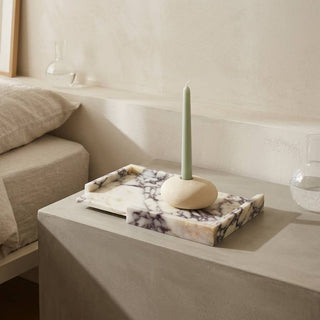 A marble candle holder tray with a light green taper candle, styled on a concrete bedside table next to a bed with neutral linens.