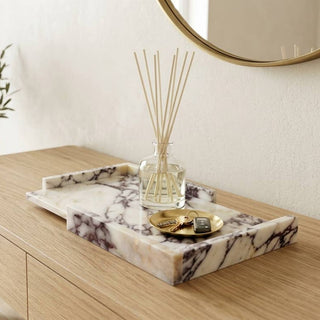 A marble tray with dark veining sits on a light wood console table in a bright, modern room. The tray holds a glass reed diffuser and a small gold dish.