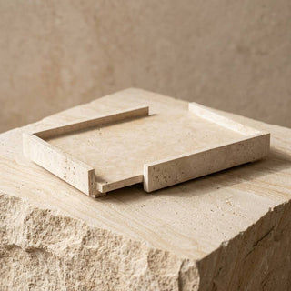 A square travertine stone tray with raised edges, shown partially slid out from its base on a matching stone pedestal against a textured beige background.