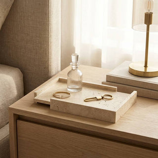 A styled bedroom scene showing a light wood nightstand beside a bed with beige bedding. On the nightstand is a rectangular stone tray holding a clear glass decanter, a gold ring, and a gold key, with books and a brass table lamp nearby.