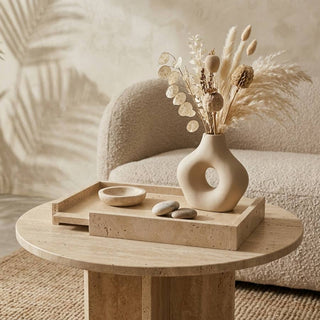 A round light wood side table in a cozy living room setting, styled with a matching wooden tray holding a beige ceramic vase with dried grasses and a small bowl.